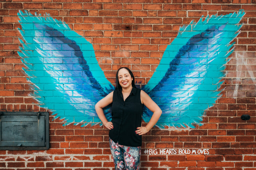 Author P.A. Cornell standing with hands on hips against a brick wall, with a turquoise wing mural behind her.